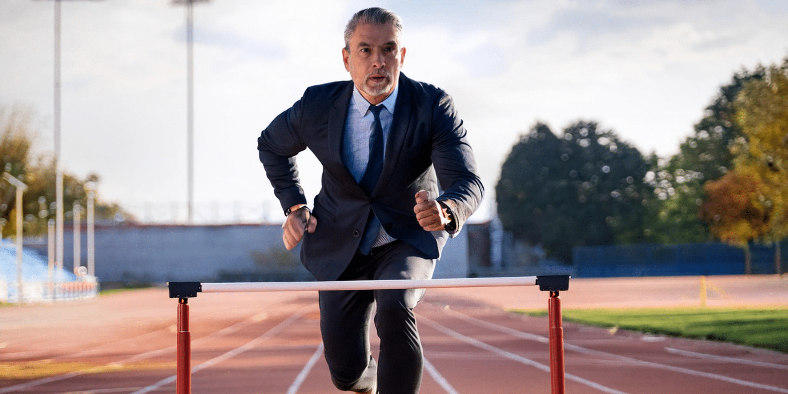 A man in his 50s wearing a business suit runs over hurdles on a track representing the obstacles in business.